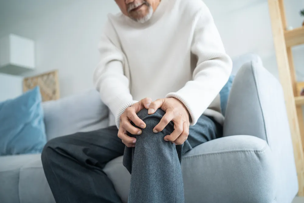 Asian Senior elderly disabled man sit on sofa suffer from injured leg. Mature older male patient sitting on couch in living room has knee problem and feel painful. Insurance and Healthcare concept.